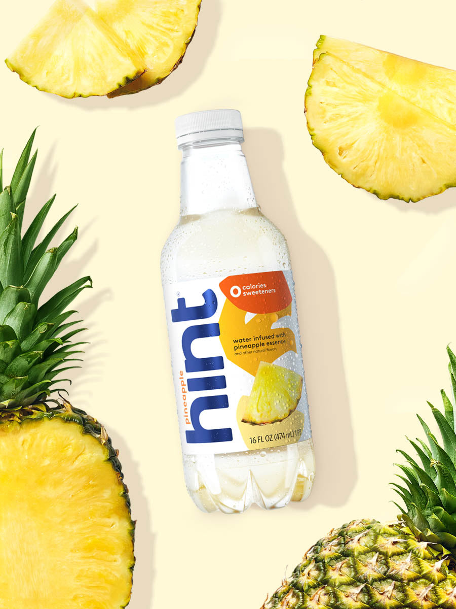 A bottle of Pineapple hint water on a yellow background. There are various pieces of fruit in the background. 