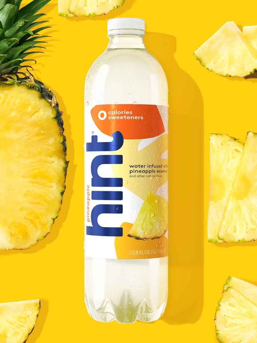 A 1L bottle of pineapple hint water on a yellow background. There are various pineapple fruit pieces around the bottle.