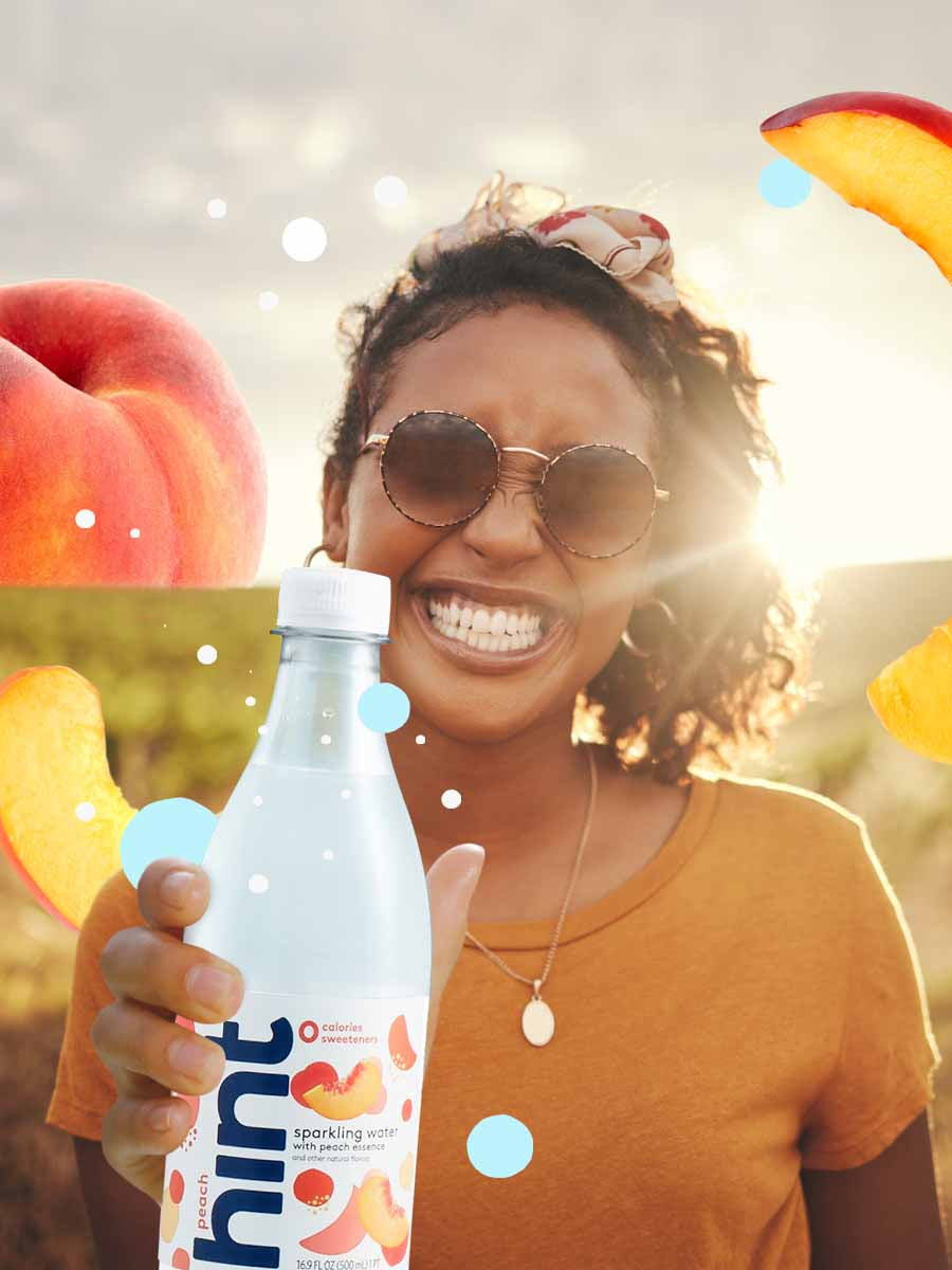 A smiling woman holding a bottle of peach sparkling hint water. The sun is setting in the background and has some peach graphics can be found in the sunset.