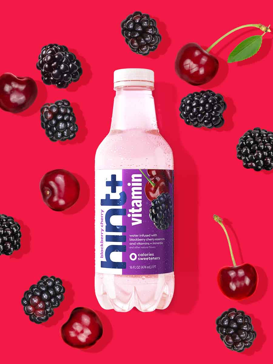 A bottle of blackberry cherry hint+ vitamin on a red background. There are various blackberry and cherry fruit pieces around the bottle.