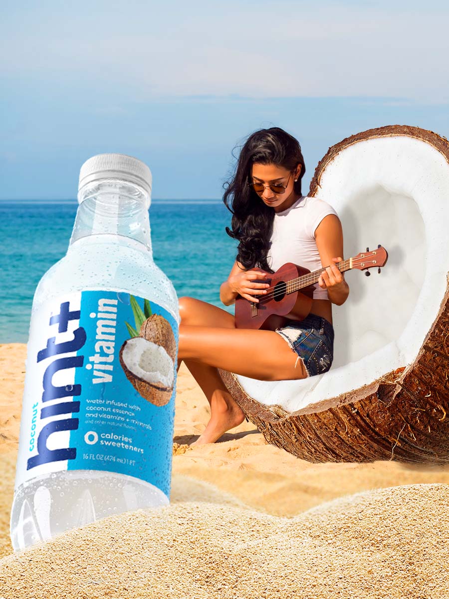 A photo of a woman sitting in an oversized chair coconut playing a ukelele at the beach. In the foreground is an oversized bottle of hint+ vitamin in the coconut flavor.