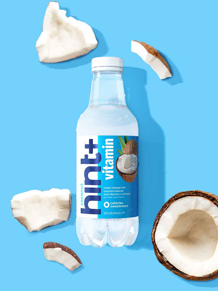 A bottle of coconut hint+ vitamin on a blue background. There are various coconut fruit pieces around the bottle.