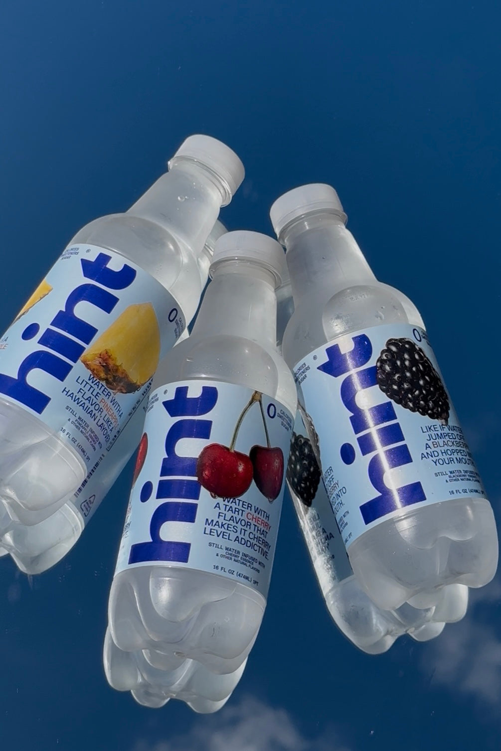 Four Hint flavored water bottles with pineapple, cherry, and blackberry labels against a blue sky background.