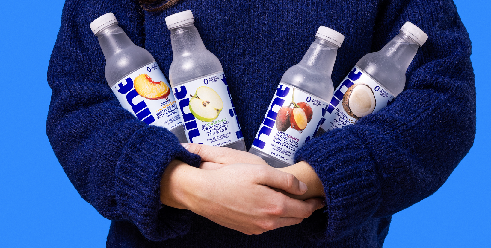 Person in blue sweater holding four Hint flavored water bottles with peach, apple, grape, and coconut images on labels