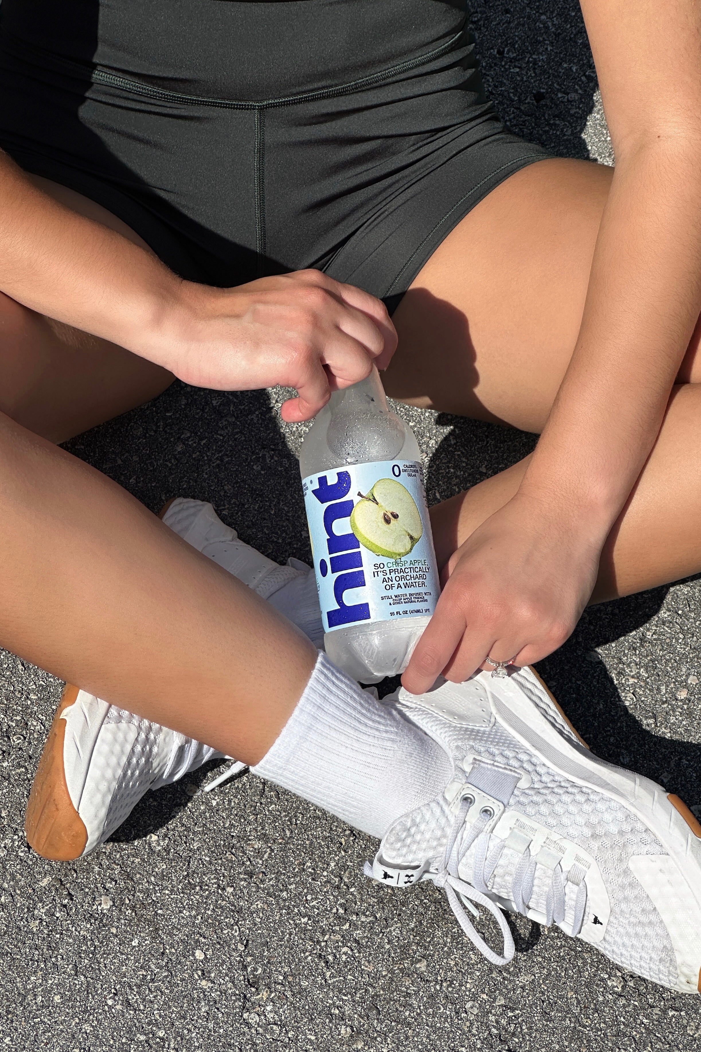 Person sitting on pavement holding a 16 fl oz bottle of Hint Water Crisp Apple flavor with visible label