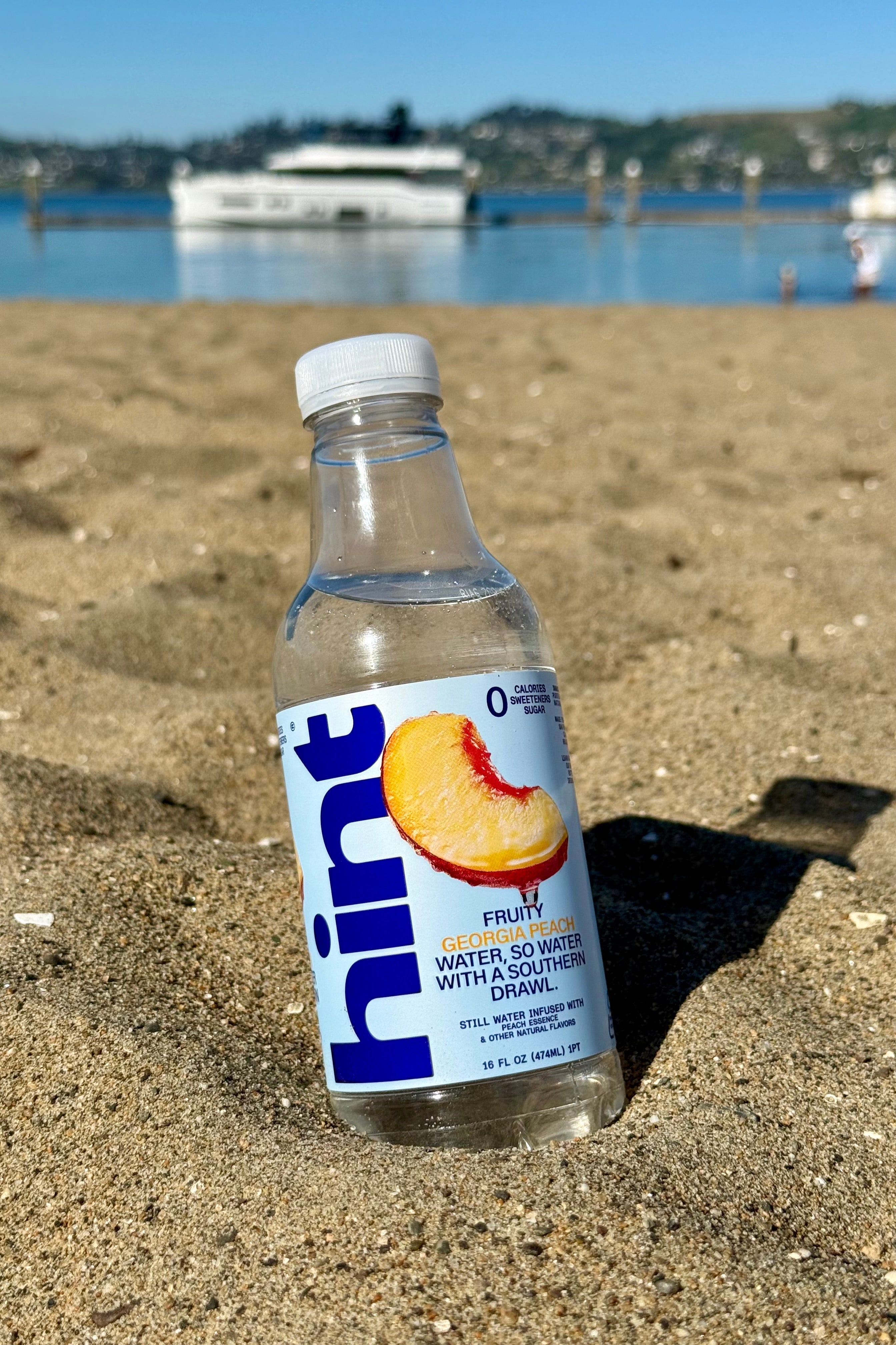16 fl oz Hint Water bottle with Georgia Peach flavor half-buried in sand on a sunny beach with water and boat in background