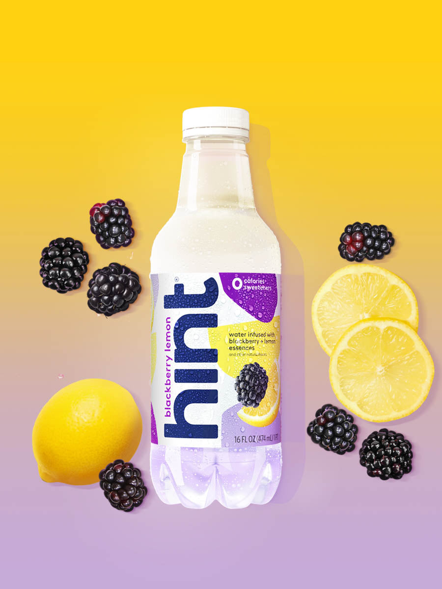 A bottle of Blackberry Lemon hint water on a purple yellow background. There are various fruit pieces in the background surrounding the bottle.