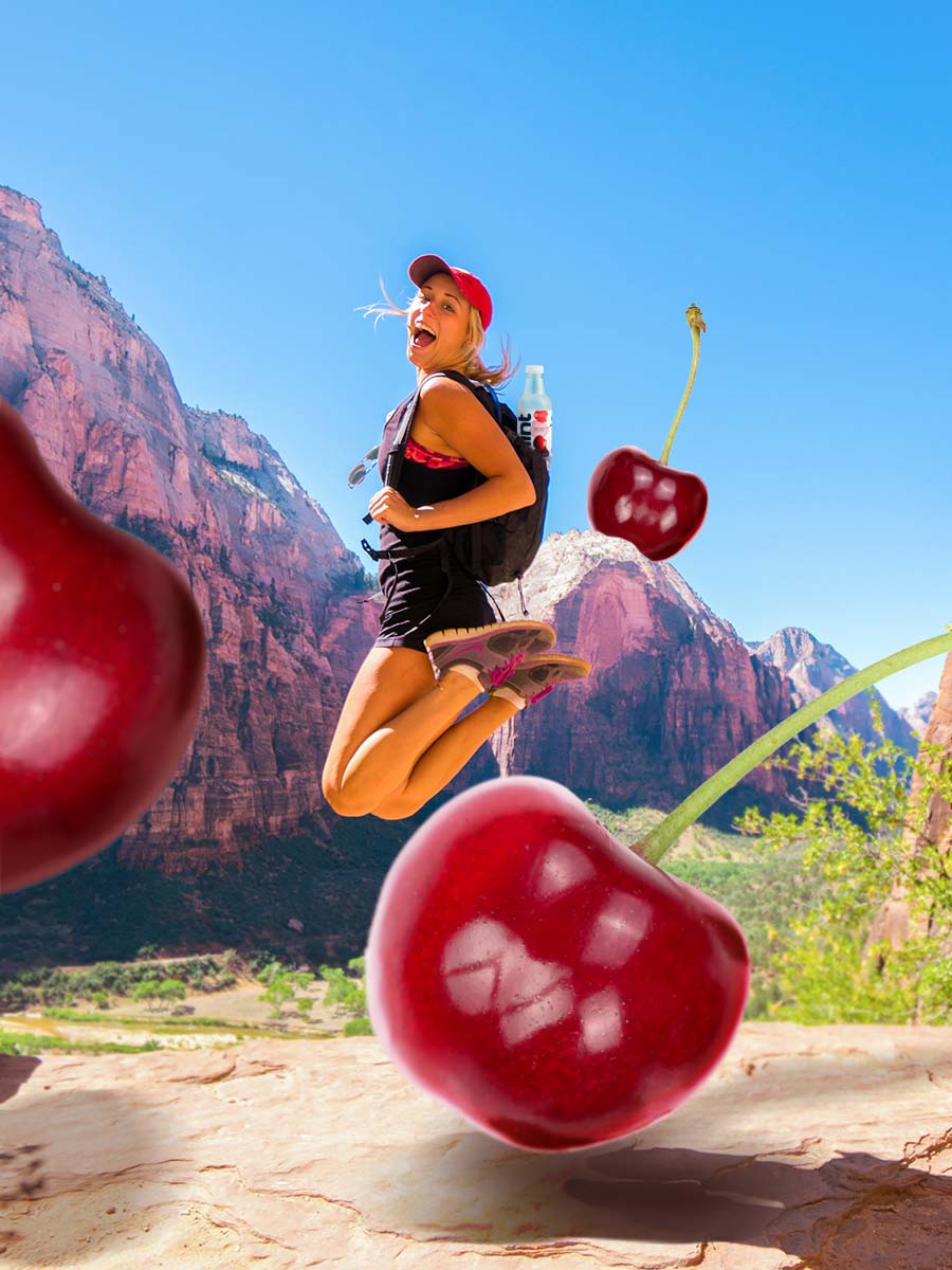 A photo of a woman jumping in the air while out on a hike with mountains in the background. There is a bottle of Cherry hint water in the backpack. There are oversized cherries in the foreground.