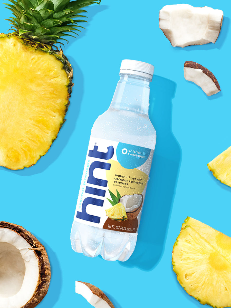 A bottle of Coconut Pineapple hint water on a blue background. There are various pieces of these fruits alongside the bottle.