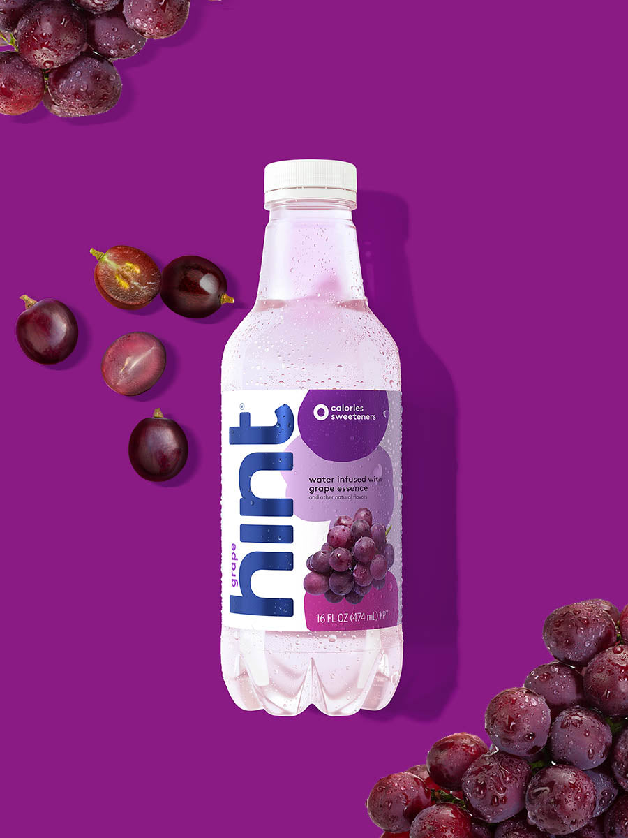 A bottle of Grape hint water on a purple background. There are various grapes alongside the bottle.