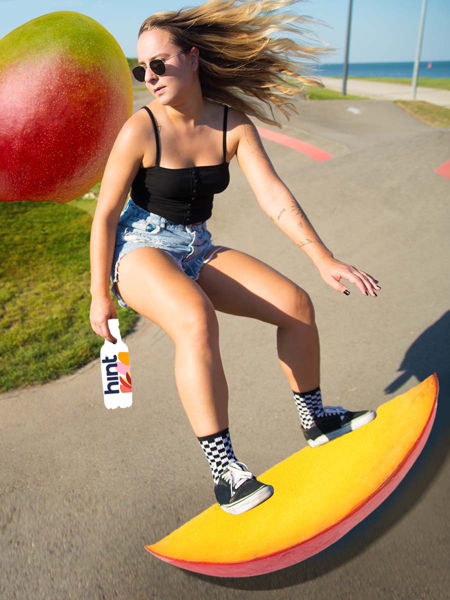 A person skateboarding on an oversized slice of mango with a bottle of Mango hint water in their hand.