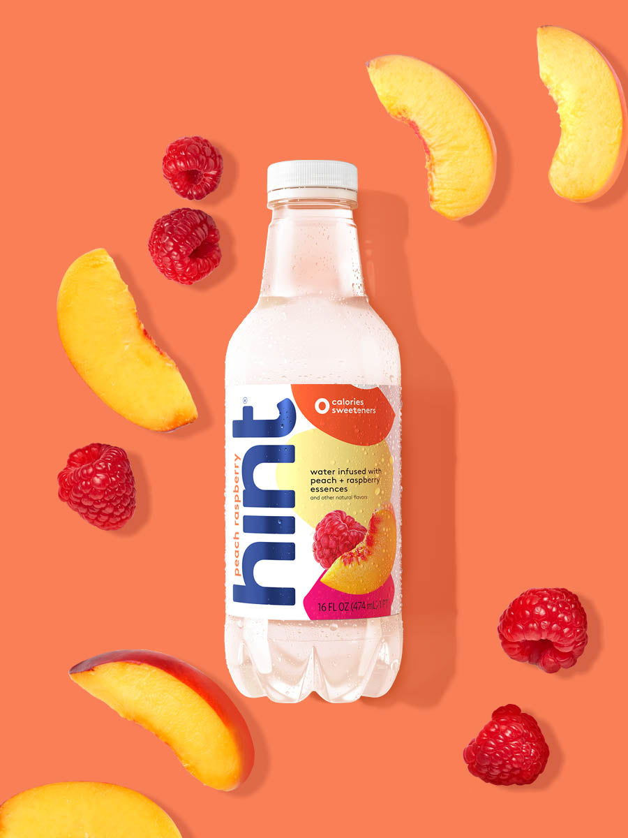 A bottle of Peach Raspberry hint water on a orange background. There are various fruit pieces in the background surrounding the bottle.