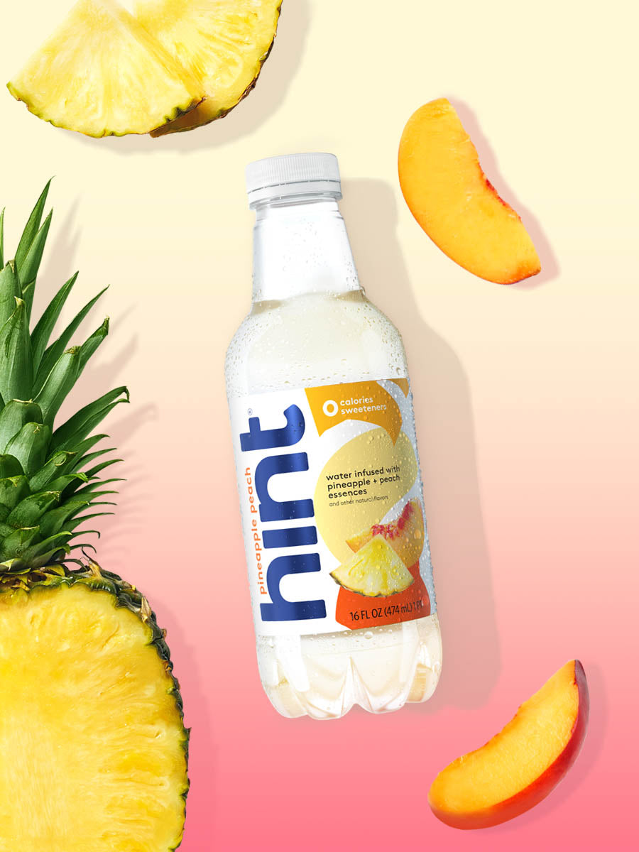 A bottle of Pineapple Peach hint water on a light pink and yellow background. There are various pieces of these fruits alongside the bottle.