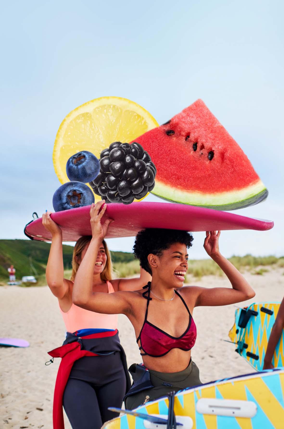 A photo of two woman carrying a surf board above their head that balances fruit graphics on top.