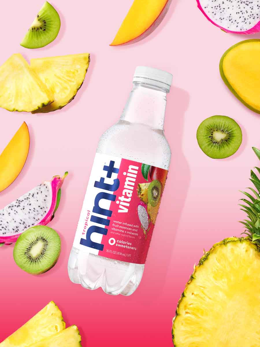 A bottle of hint+ vitamin in the tropical flavor on a gradient pink background. There are cutout images of fruit in the background.