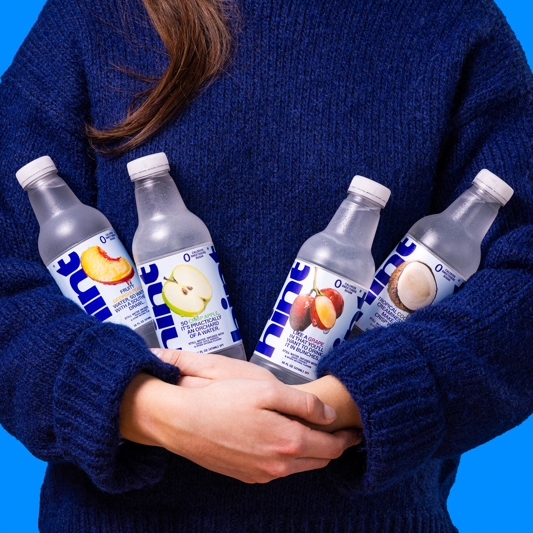 Person in blue sweater holding four Hint flavored water bottles with peach, apple, grape, and coconut flavors
