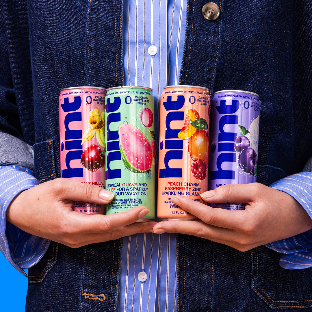 Four cans of Hint sparkling water flavors held in hands: Pineapple Vanilla, Tropical Guava Lychee, Peach Charm Raspberry, Wild Berry.