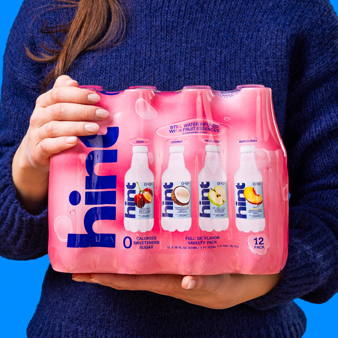 Person holding a 12-pack of Hint flavored still water in grape, coconut, crisp apple, and Georgia peach varieties