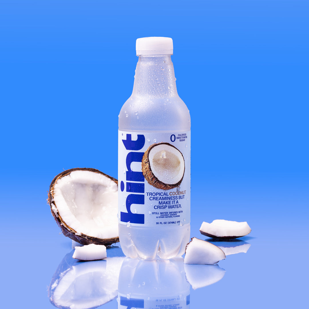 Bottle of Hint coconut flavored water with coconut halves and slices on reflective blue surface