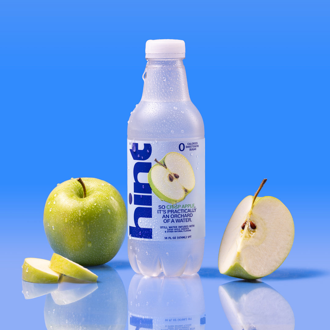 Clear plastic bottle of Hint water with green apple flavor, next to whole and sliced green apples against blue background.