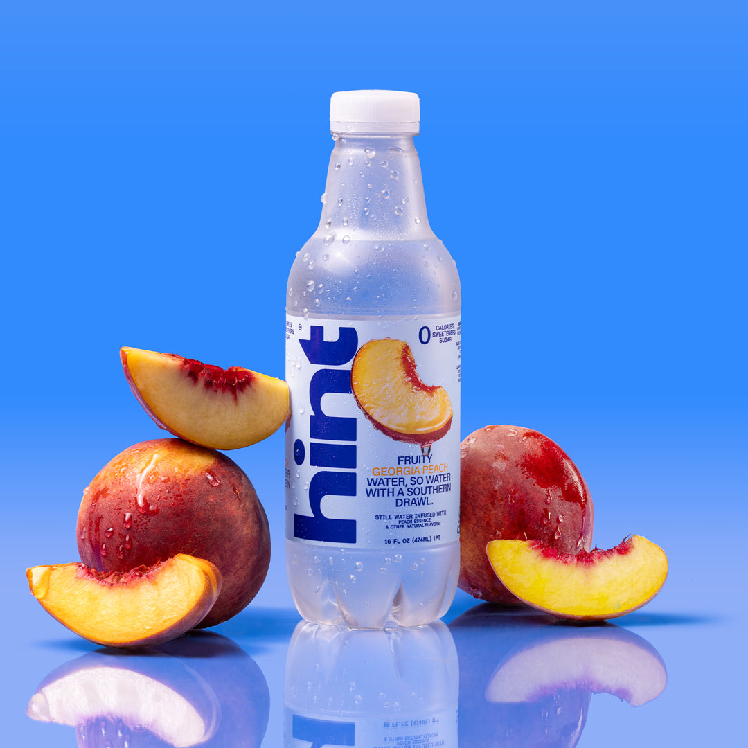Bottled Hint flavored water with Georgia peach essence surrounded by fresh peach halves on a blue gradient background