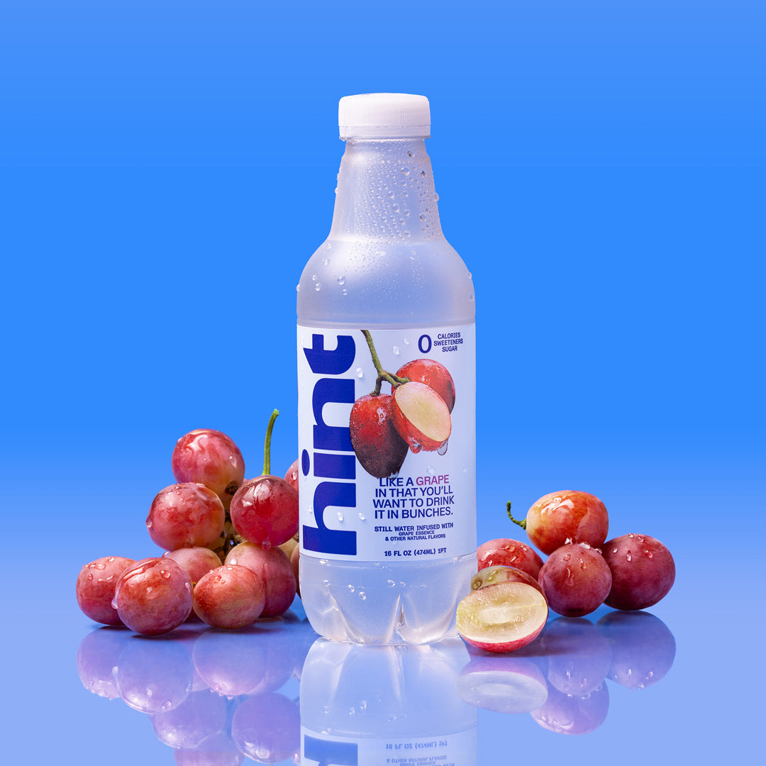 Plastic bottle of Hint grape flavored water surrounded by red grapes on reflective surface