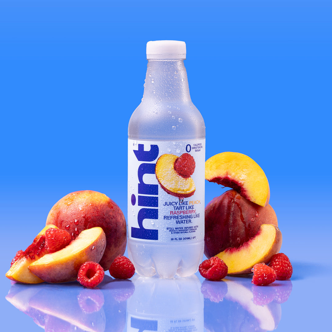 Clear plastic bottle of Hint flavored water with peaches and raspberries around it on reflective blue surface