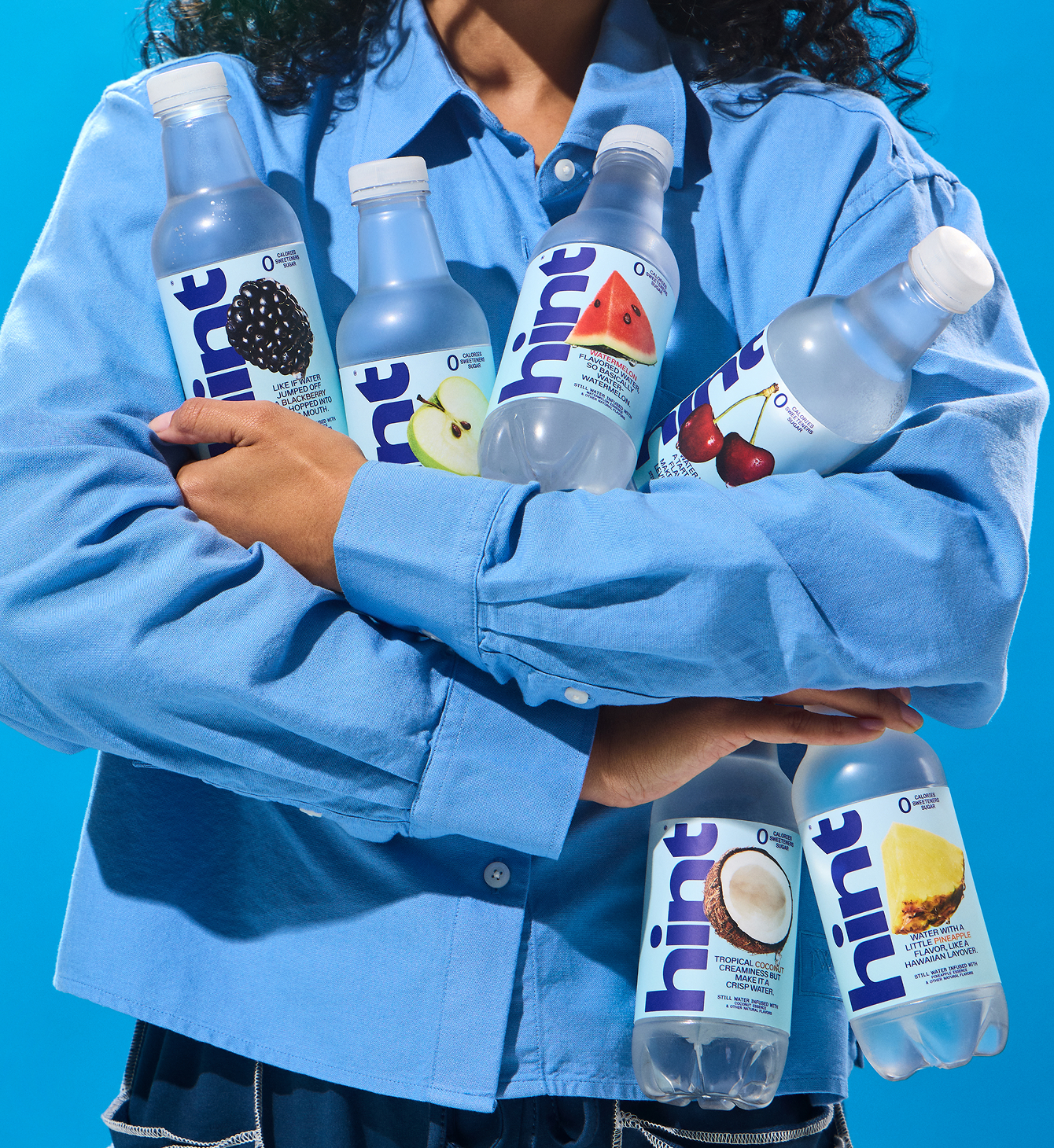 Person in blue shirt holding six Hint flavored water bottles with blackberry, green apple, watermelon, cherry, coconut, and pineapple labels